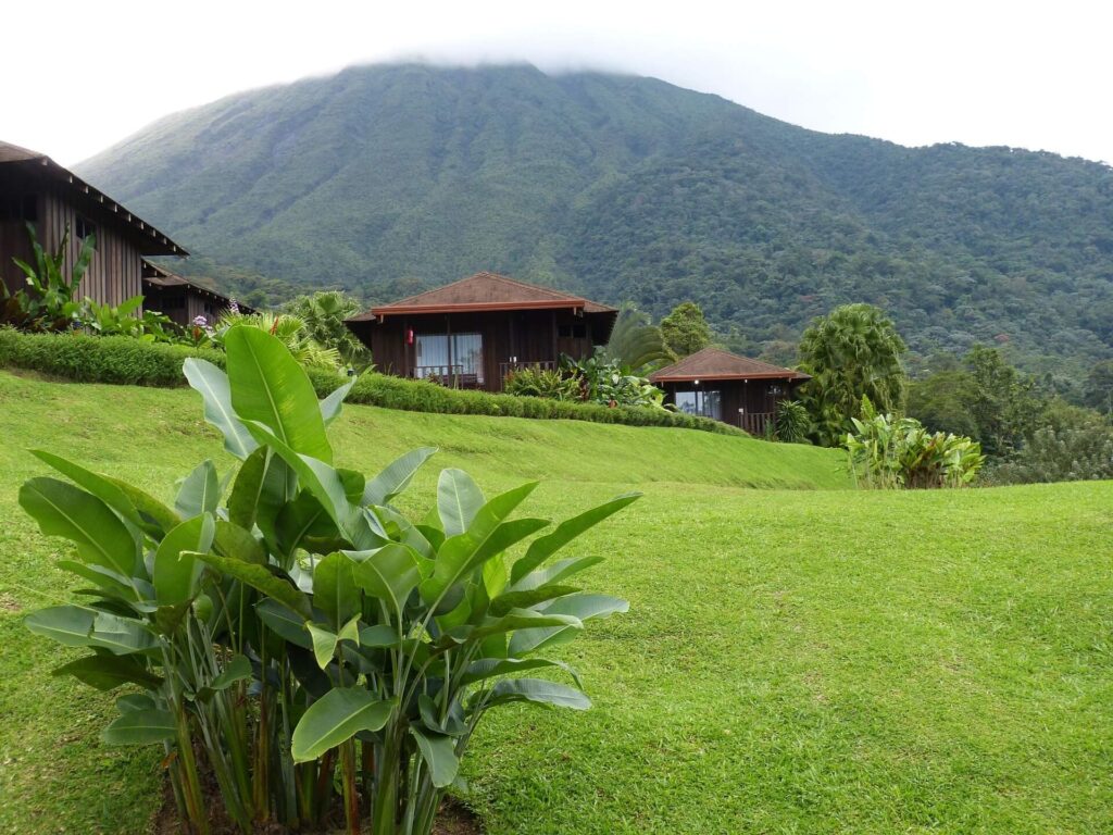 Wooden eco-lodge bungalows on a green hillside with a mist-covered volcanic peak rising behind lush tropical forest, similar to lodge accommodations near Costa Rica's volcanic highlands