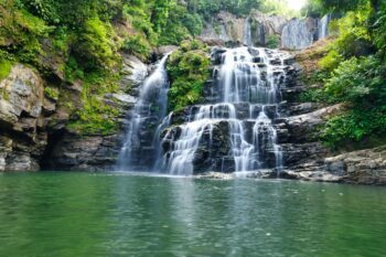 Multi-tiered waterfall cascading over moss-covered rocks into an emerald green swimming pool surrounded by lush Costa Rica rainforest