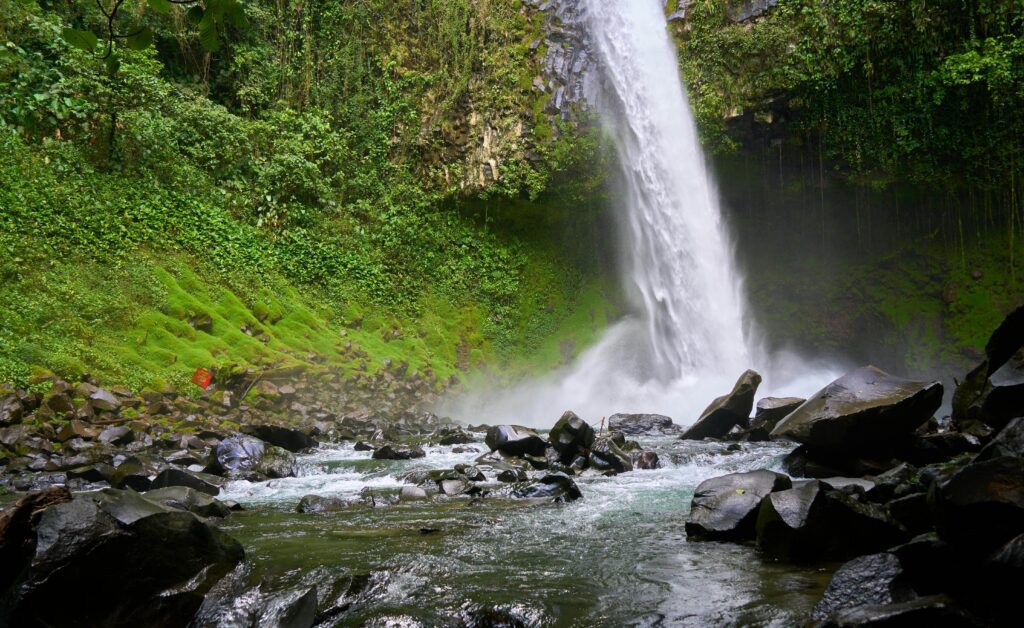 Powerful waterfall plunging over a vine-covered cliff onto large boulders with a rocky stream flowing through dense green vegetation in Costa Rica