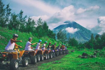 Group of ATV riders lined up on a green hillside with a cloud-wrapped volcano rising in the background, similar to views near Arenal Volcano in Costa Rica