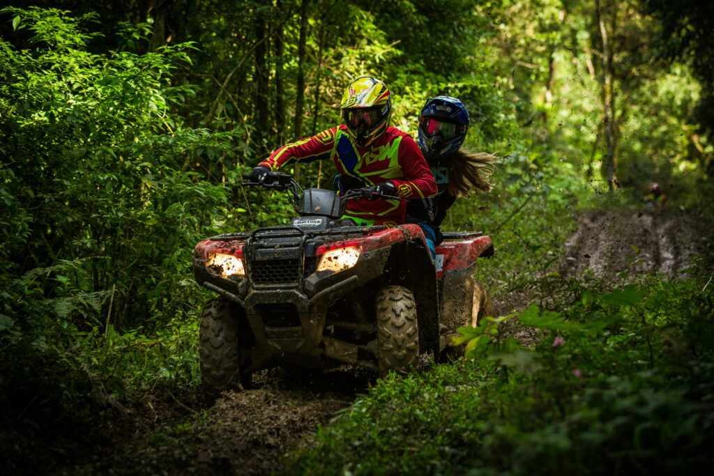 Two riders in full motocross gear powering a mud-covered ATV through a narrow jungle trail during Costa Rica's green season