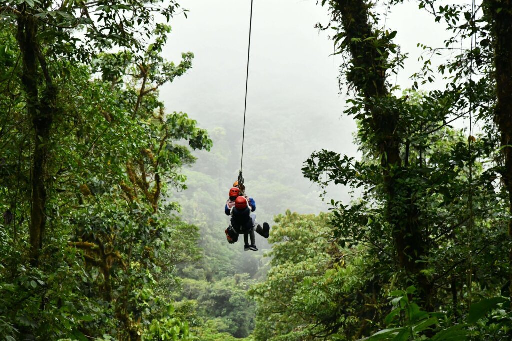 Two visitors in safety helmets riding a zipline through lush Costa Rican rainforest canopy, similar to the 8-cable canopy tour available at Hacienda La Chimba near San José