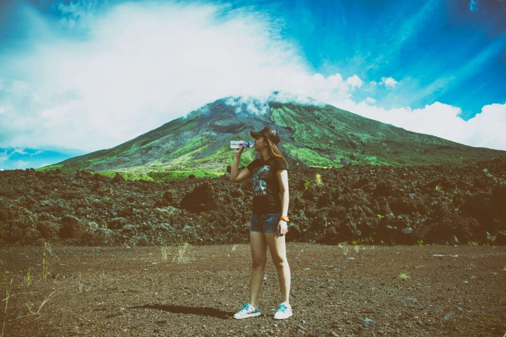 A traveler drinking water while standing on a volcanic lava field in front of a green-flanked Costa Rica volcano under a bright blue sky, showing the rugged terrain typical of volcanic hiking areas