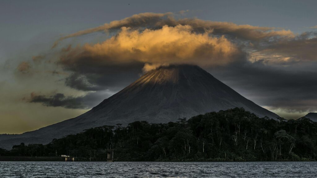 A conical Costa Rica volcano rising above a lake at sunset, surrounded by dense tropical forest, with dramatic golden clouds wrapping the summit against a dark evening sky