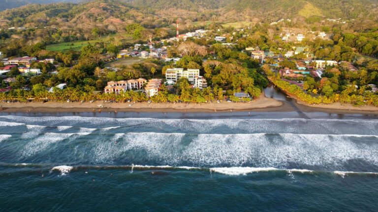 Aerial view of Jaco, Costa Rica, showing the beachfront, waves, and town backed by green hills.