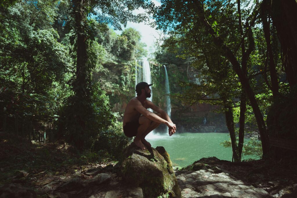 Visitor sitting on a mossy rock overlooking a waterfall and swimming hole similar to La Fortuna Waterfall in Costa Rica