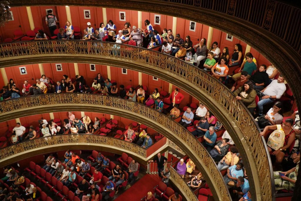 Packed audience seated across multiple ornate balcony tiers inside Teatro Nacional, Costa Rica, with gold ironwork railings and red seating