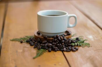 White ceramic cup of freshly brewed Costa Rican coffee surrounded by roasted coffee beans and green fern leaves on a wooden table at a coffee farm