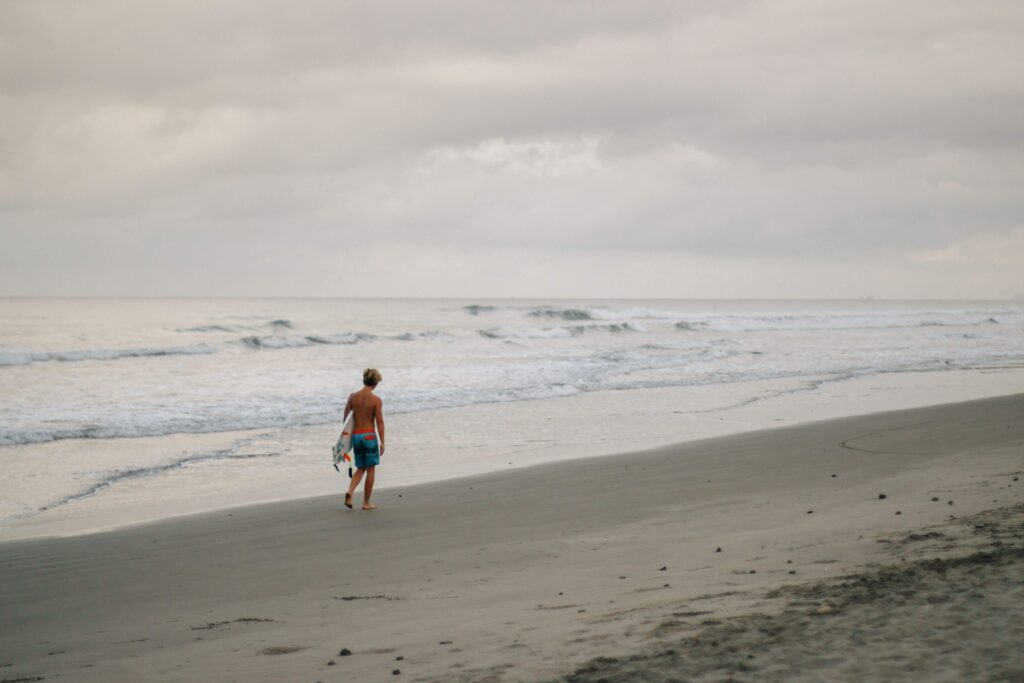 Surfer walking along an empty dark-sand beach carrying a board under an overcast sky, similar to a quiet Costa Rica surf beach