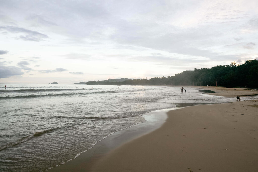 Calm shallow water and wide sand at dawn with a few swimmers and a forested headland, similar to Playa Cabuyal, Costa Rica