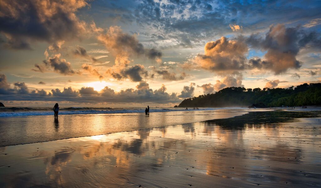 Golden sunset reflecting on wet sand as people wade near the shore, similar to a Pacific sunset at Playa Hermosa, Costa Rica